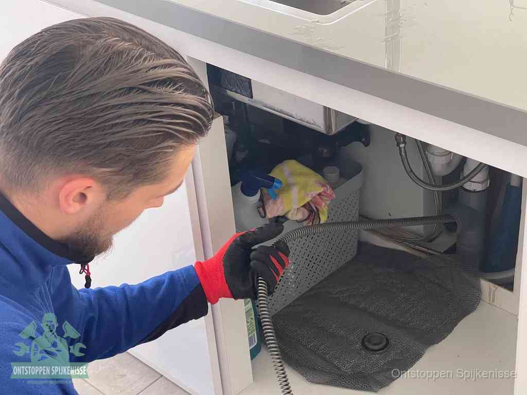 Een monteur voert een flexibele ontstoppingsveer in onder een keukenkastje om een verstopping voorbij de sifon te bereiken in Spijkenisse